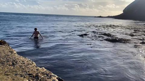 Shaun Kelly is standing waist deep in the sea, the sun is low in the sky. Mr Kelly is to the left, with rocks in the foreground in front of him, and a sillhouetted coastline to the right - it is a calm day.