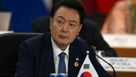 South Korea's President Yoon Suk Yeol sits in a meeting in a dark blue suit behind a South Korean flag