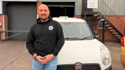 A man with short hair and beard, wearing a black hoodie and blue jeans. He is perched on the bonnet of a white fiat van.