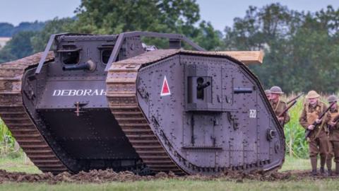 A full sized replica of a tank is pictured in a muddy field with some reenactors standing behind it wearing WW1 uniforms