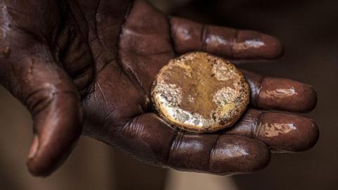 A close-up of a mud-stained hand holding a roughly circular gold nugget
