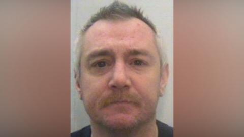 The mugshot of David Jack, a man with light grey hair and a light moustache with stubble. He is starting impassively forwards. 
