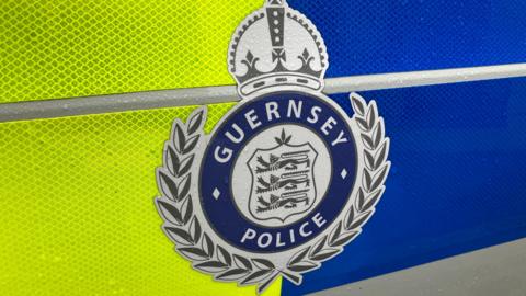 The side of a yellow and blue police vehicle. At the centre, on the door, is a Guernsey Police badge. 