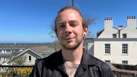 Josh Coutu pictured brunette hair tied back. He has a dark, short, beard and moustache. Josh is wearing a black button down shirt and is smiling with closed lips at the camera.