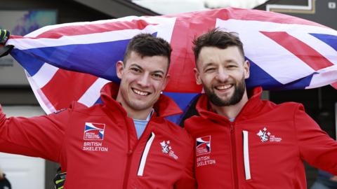 Matt Weston (left) and Marcus Wyatt stand with their arms around each other while holding a Great Britain flag in the other hand each behind them