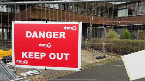 A side entrance to County Hall is blocked by metal fencing and a red sign reading 'Danger Keep Out'. Behind it are slightly overgrown flowerbeds and bushes in front of red-brick walls.