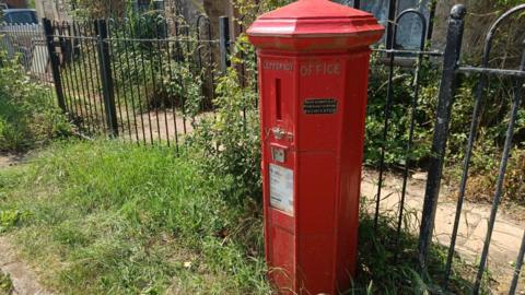 An octagon shaped red UK postbox is nestled in long grass in front of black metal fencing.