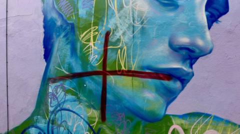 A dark red cross painted on top of a blue and green mural of a man looking down. It is painted on a purple wall, and there are several other blue and cream scribbles - which appear to be part of the mural - over his face.