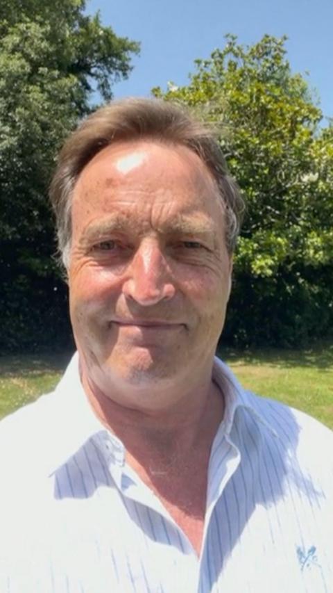 A man with brown hair in a white top. Behind him is green grass and a green tree.