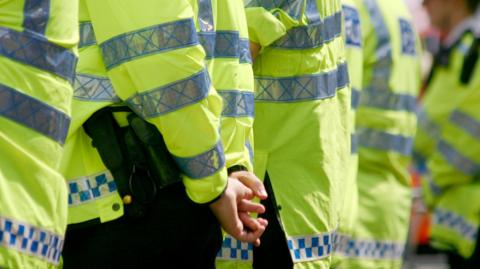 A row of policemen controlling a crowd. Focus is on hands and body of left-most policeman who has his hands interlaced behind his back. The officers are all wearing bright yellow high-vis long-sleeved jackets with silver reflective panelling on them and blue and white check strips along the hem.