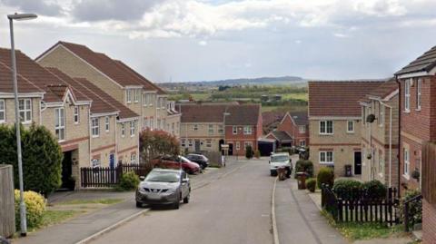 A residential street in Barnsley, with the road a downward slope into the distance. The homes on both sides of the road are relatively new. A hill with trees can be seen in the distance. 