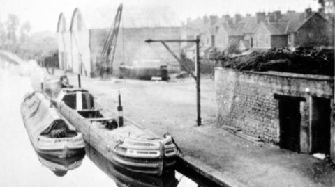 A black and white photo of Melksham Wharf taken around 1900. There are two canal boats on the water. A number of buildings and a crane can be seen on the bank.