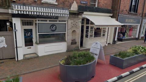 Two store fronts with a historic town wall gate post in between them. The shops are fronted by planters on an extended pavement. An A-board advertising the deli sits in front of the shop. 