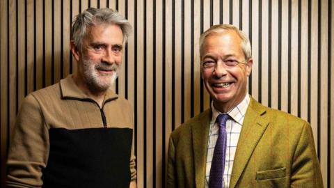 Christopher Harborne, left, wears a beige and black jumper. He has a full head of grey hair and a grey beard and bushy eyebrows. He stands next to Farage who is grinning. Farage is wearing a green suit and checked shirt and purple tie. 
