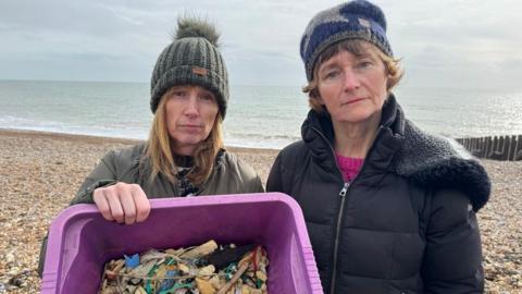 Lisa Laing is wearing a green jacket and bobble hat, and Nicola Lambert is wearing a black outdoor coat and navy hat. Laing is holding a purple box of debris that has been picked up from the shingle beach. They are standing with the sea behind them.