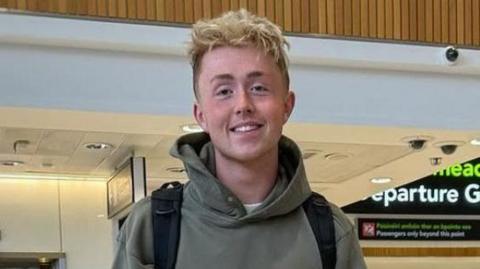 Jamie Harkin is standing in the airport departures area. He is wearing a green hoodie and wearing a black bag pack. There is an elevator behind him with two people standing outside of it. To his side there are four people with two hugging. In the background there is a large black departures gate.  