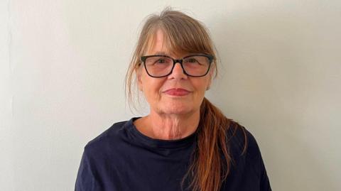 A woman is stood against a white wall looking directly at the camera. She is wearing black glasses and a navy blue shirt. 