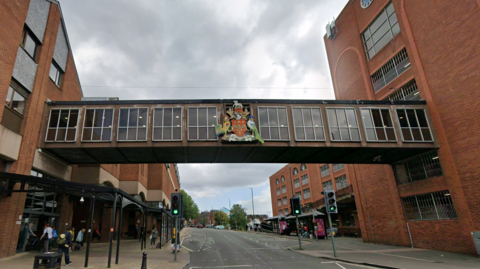 A footbridge between two brick buildings spanning a street bearing a civic crest 