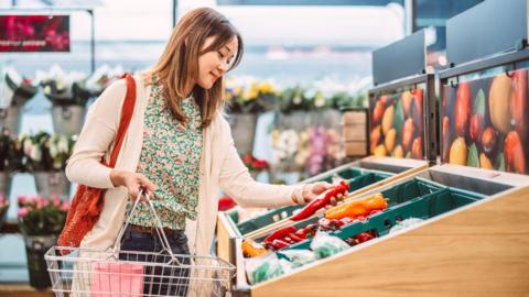 A woman holds a pepper in the supermarket as she shops holding a basket