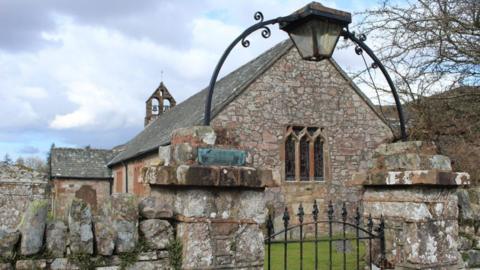 A stone wall with black iron gate below an ornate lamp hung from a metal arch. An old stone church with an open bell tower sits behind. 