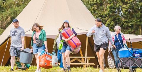 A group of people walk across a field carrying and pulling camping equipment. There are four woman and two men in a line walking in front of a teepee-style tent