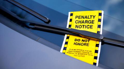 A bright yellow parking fine is tucked under a car's window wiper