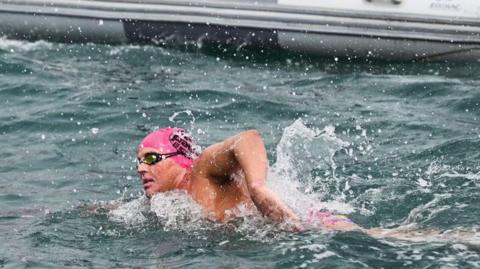 William Ellis is swimming in blueish-grey sea besides a grey inflatable support boat. He has a pink swimming cap and is wearing goggles.