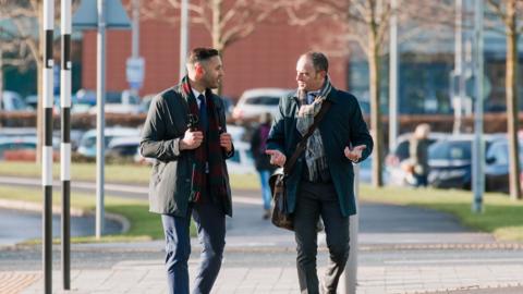 Two businessmen walk to work talking to each other.
