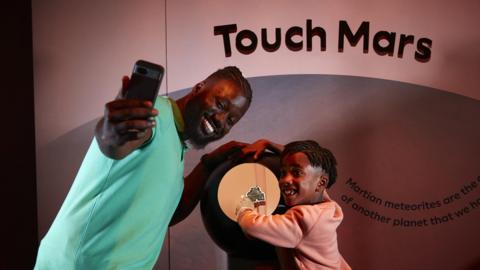 A man and a child pose for a selfie with an iPhone in front of a meteorite from Mars.