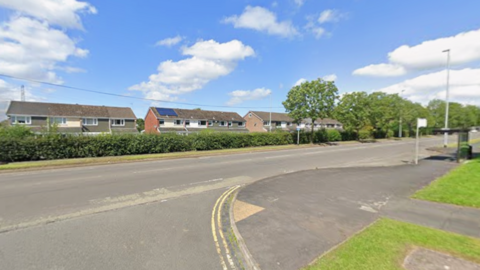 The junction of two roads, with several houses and bushes on the furthest side of the road.