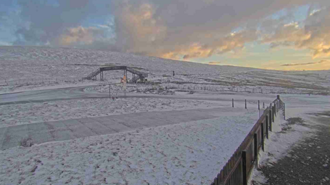 A webcam view of the road at the Bungalow looking towards Onchan. There is a metal footbridge in the distance and the ground is covered with snow.