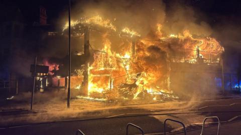 A fire is earing through a two storey building and has burnt down the walls, just leaving the bones and foundations of the site. It remains engulfed in flames. The sky is dark making the fire appear even brighter against the night sky.