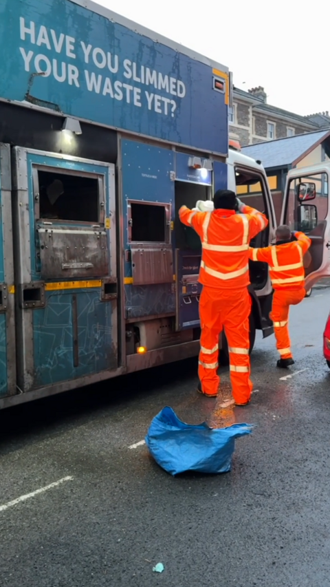 Two bin collection workers dressed in orange hi-vis jackets and trousers, emptying recycling bins into the side of a rubbish truck in the middle of the road.