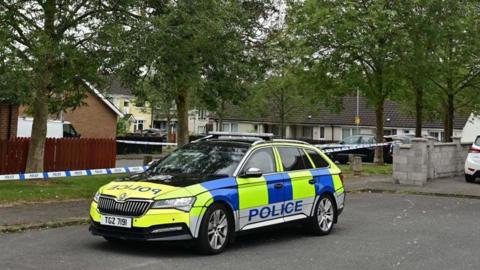 A police car which is yellow and blue in colour is parked alongside police cordon tape. Police is written on the side in blue letters. There are houses and trees in the background.