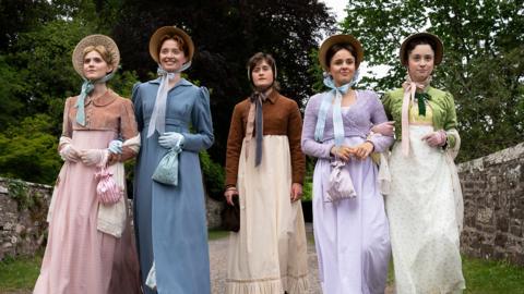 Five individuals walking side by side along a tree‑lined path bordered by a stone wall. They are all dressed in Regency‑style clothing,