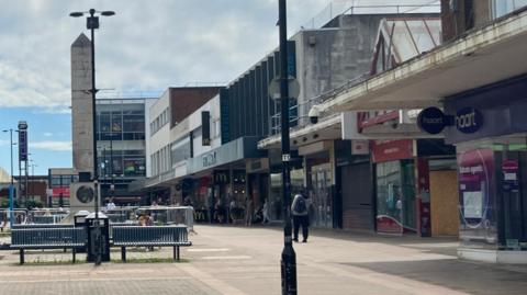 A shopping centre with benches in the middle and shops on the side, street lamp in the corner and people walking down the high street in broad daylight