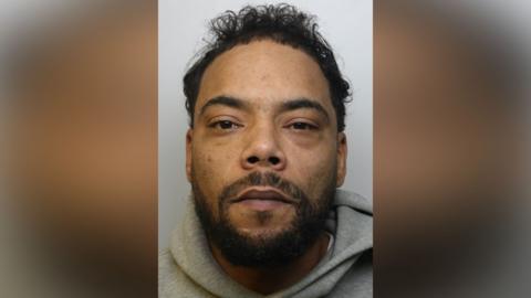 Photo of Jamie Ivers' prison mug shot. He is looking directly into the camera. The headshot photo includes a small portion of his grey hoodie. Jamie has black hair and a beard. 