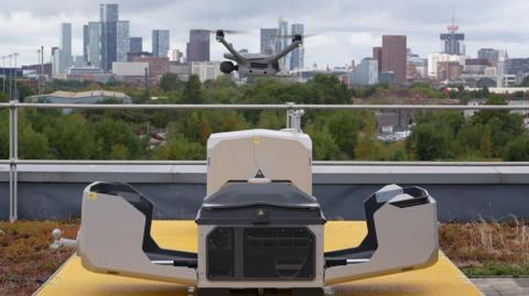 A drone hovering above an open docking box, with the Manchester skyline in the background.