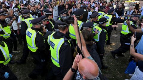 Police officers detain a protester during a rally aimed at challenging the government's proscription of Palestine Action under anti-terrorism laws, in in London, Britain on 9 August. The picture shows a cluster of police in high-vis jackets, holding a man, with a crowd surrounding them. In the foreground, a man takes pictures with a camera.