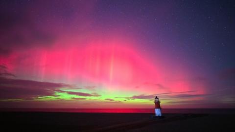 Landscape shot of the northern lights from the Point of Ayre. A lighthouse can be seen looking over the coast whilst the sky is lit in bright pinks, greens and purple.