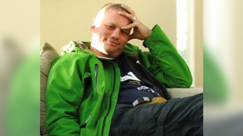 Mark Pell sitting on a couch reclining. He's wearing a bright green waterproof jacket open at the front and is smiling with one hand on top of his head and forehead 