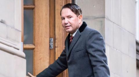 A smartly dressed man, with dark hair swept to one side, enters a building wearing a grey coat.