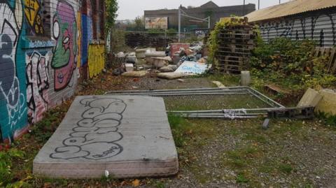 A mattress, sofas and bags of rubbish discarded next to a wall which has been graffitied.