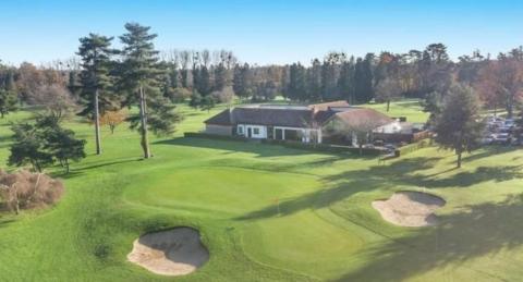 One of the putting greens and two bunkers on the golf course, which is dotted with fir trees. In the middle of the photo is a single storey building surrounded by a low hedge, with a car park next to it.