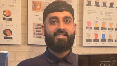 Close up of Usman Hussain in the cafe, he has short dark hair and a beard and is wearing a navy polo shirt