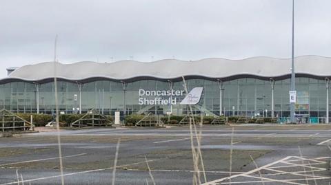 Exterior of Doncaster Sheffield Airport. It has a wavy roof and the front is fully glass. The car park in the foreground is empty.