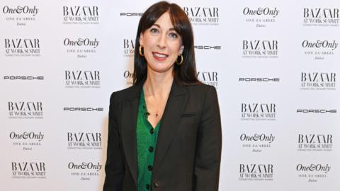 Samanthan Cameron wearing a green cardigan under a black blazer, at a magazine event with the promotional board behind her. She is smiling and wearing gold hoop earrings.