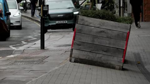 The sloping pavement with a big planter on it. The background includes cars and pedestrians.