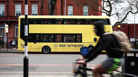 A yellow Bee Network bus drives down Oxford Road in Manchester. Nearer the camera, a deliberately out-of-focus male cyclist rides along a cycle path.