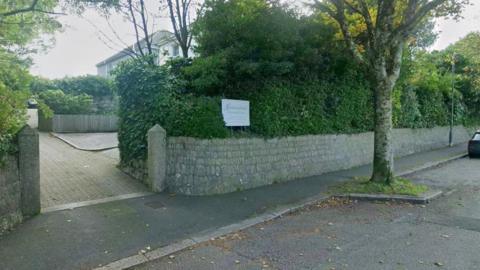 A picture of the front of Garsewednack Residential Home. There is a large stone wall with trees along the road. There is a small entrance into the home grounds next to a sign.